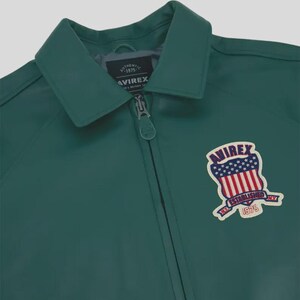 Puede incluir: Chaqueta verde oscuro con cuello y cierre de cremallera. La chaqueta presenta un parche con el texto "AVIREX ESTABLISHED NY 1975" en rojo, blanco y azul. La chaqueta es de material liso.
