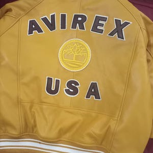 May include: Mustard yellow leather jacket with the word "AVIREX" and "USA" in brown lettering. A circular patch with a tree design is centered below the "AVIREX" text. The jacket has a white and tan striped band at the waist.