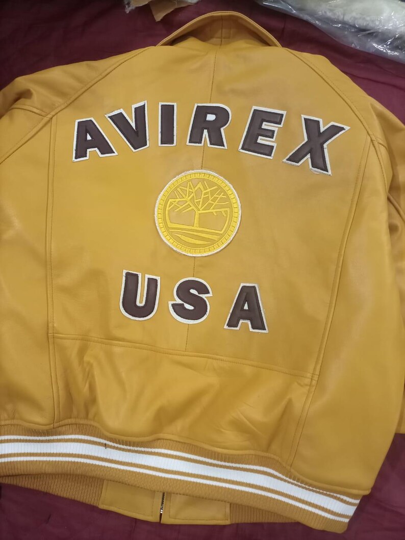 May include: Mustard yellow leather jacket with the word "AVIREX" and "USA" in brown lettering. A circular patch with a tree design is centered below the "AVIREX" text. The jacket has a white and tan striped band at the waist.