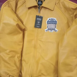 May include: Mustard yellow leather jacket with a zip-up front, collar, and a patch on the chest. The patch features a shield design with stars and stripes, and the text "AVIREX ESTABLISHED 1975". The jacket has white and yellow striped cuffs.