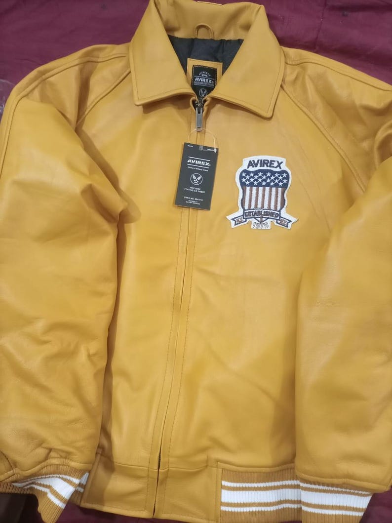 May include: Mustard yellow leather jacket with a zip-up front, collar, and a patch on the chest. The patch features a shield design with stars and stripes, and the text "AVIREX ESTABLISHED 1975". The jacket has white and yellow striped cuffs.