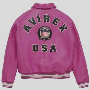 Könnte beinhalten: Eine leuchtend pinkfarbene Lederjacke mit dem Wort "AVIREX" in schwarzer Schrift auf dem Rücken. Darunter befindet sich ein kreisförmiger Aufnäher mit der Aufschrift "ALL AMERICANS ESTABLISHED 1975 USA". Die Jacke hat einen weiß-schwarz gestreiften Besatz.