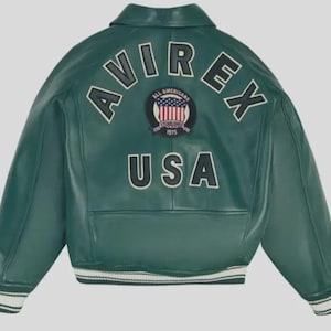 Puede incluir: Chaqueta de cuero verde con ribetes blancos en los pu&ntilde;os y el dobladillo. La parte trasera de la chaqueta presenta la palabra "AVIREX" en letras blancas en bloque sobre un parche circular con las palabras "ALL AMERICANS ESTABLISHED 1975" y la bandera de EE. UU. Debajo, la palabra "USA".