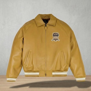 May include: Mustard yellow leather bomber jacket with a collared neckline, zipper closure, and ribbed cuffs and hem. Features a patch on the chest with the text "AVIREX 1975". The jacket has two front pockets and a classic bomber silhouette.