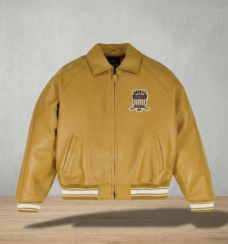 May include: Mustard yellow leather bomber jacket with a collared neckline, zipper closure, and ribbed cuffs and hem. Features a patch on the chest with the text "AVIREX 1975". The jacket has two front pockets and a classic bomber silhouette.