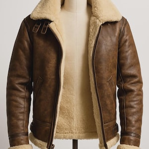 May include: A brown leather jacket with a cream shearling lining and collar. The jacket has a zip-up front, two front pockets, and a buckled collar. The jacket is displayed on a mannequin.