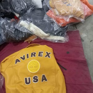 May include: Mustard yellow leather jacket with the word "AVIREX" across the back in brown lettering. Below the brand name is a circular patch with a tree design and the word "USA". Other jackets are visible in the background.