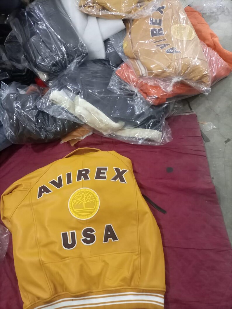 May include: Mustard yellow leather jacket with the word "AVIREX" across the back in brown lettering. Below the brand name is a circular patch with a tree design and the word "USA". Other jackets are visible in the background.