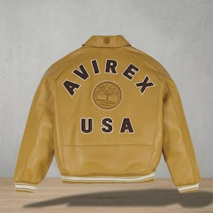 May include: Mustard-colored leather jacket with the word "AVIREX" arched across the back, above "USA". A circular emblem is centered between the words. The jacket has a collar, ribbed cuffs, and a ribbed hem with white stripes.