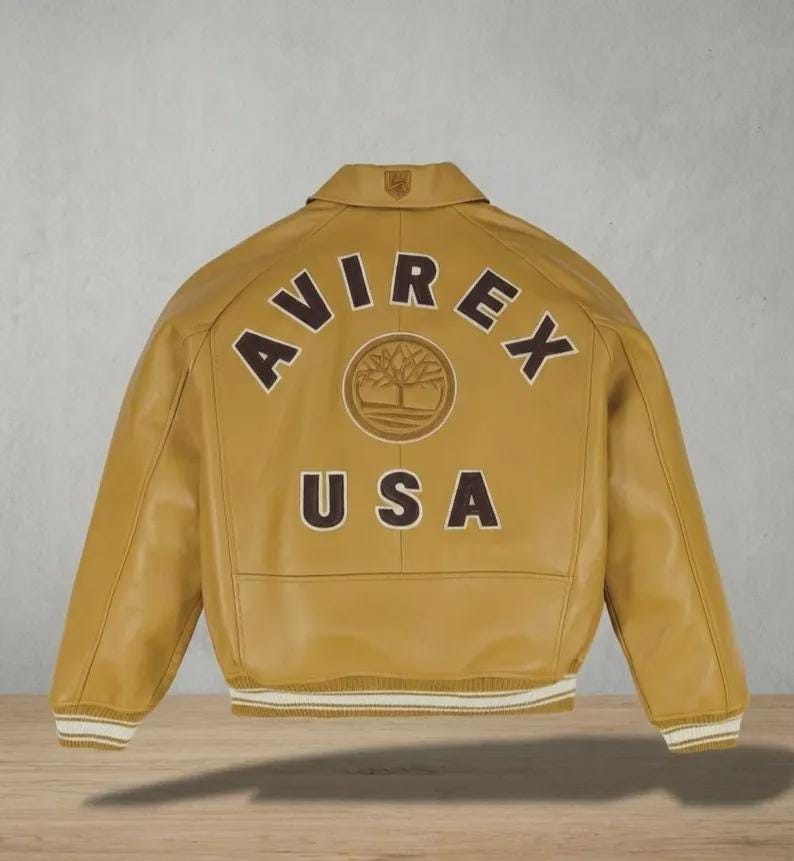 May include: Mustard-colored leather jacket with the word "AVIREX" arched across the back, above "USA". A circular emblem is centered between the words. The jacket has a collar, ribbed cuffs, and a ribbed hem with white stripes.