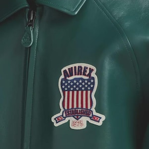 Puede incluir: Primer plano de una chaqueta de cuero verde oscuro con cremallera y un parche. El parche muestra la palabra "AVIREX" sobre un dise&ntilde;o de la bandera estadounidense y las palabras "ESTABLISHED 1975 NY".