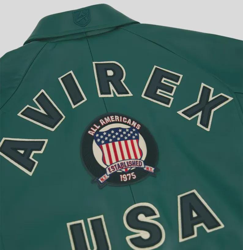 Puede incluir: Chaqueta verde oscuro con la palabra "AVIREX" en letras blancas con contorno azul oscuro. Un parche circular con la bandera americana y las palabras "ALL AMERICANS ESTABLISHED NY 1975" est&aacute; centrado debajo.