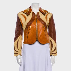 Peut inclure: Veste en cuir multicolore avec fermeture éclair et deux poches zippées. Le design de la veste est composé de blocs de couleurs marron, beige et orange. Le col et les manches présentent des découpes décoratives. La veste est présentée sur un mannequin.