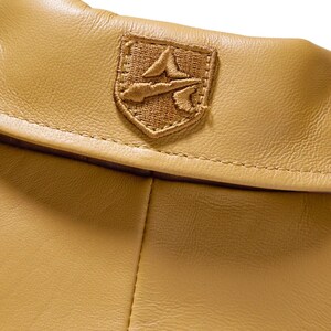 May include: Close-up of a tan leather garment with a stitched emblem. The emblem is a shield shape with a detailed embroidered design in a matching tan color. The leather has a subtle texture.