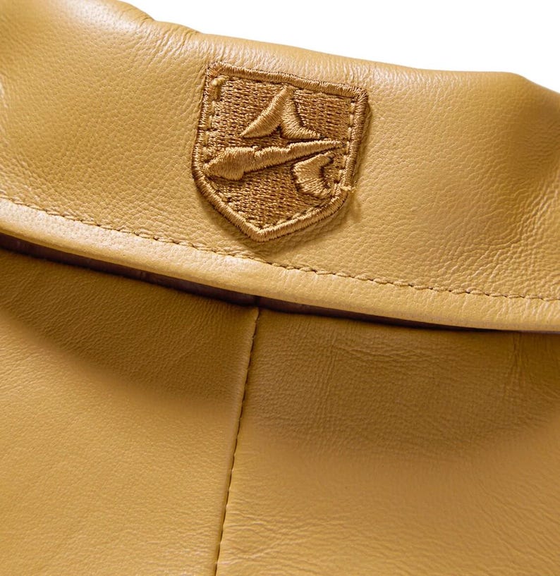 May include: Close-up of a tan leather garment with a stitched emblem. The emblem is a shield shape with a detailed embroidered design in a matching tan color. The leather has a subtle texture.