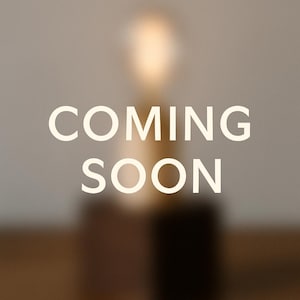 May include: A blurred image with the words "COMING SOON" in white, centered. The background features a light fixture with a warm glow, and a dark base. The overall aesthetic is minimalist and modern.