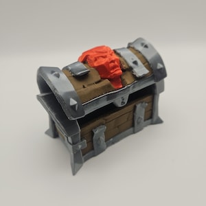 May include: A small treasure chest model. It features a brown wooden body, grey metal detailing, and a red sculpted element on the lid. The chest has a hinged lid and is designed with a medieval aesthetic.