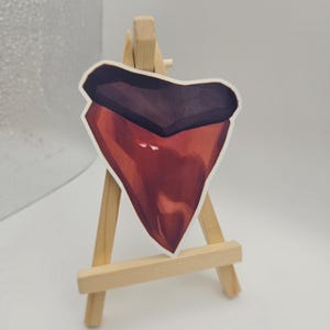 May include: A sticker of a shark tooth, with a dark brown top and a gradient of red to light red. The sticker is displayed on a small wooden easel against a white background.