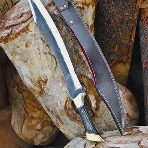 Puede incluir: Una espada artesanal con una hoja larga y curvada y un mango negro con detalles dorados. La espada se muestra con una funda de cuero marrón con forro rojo, sobre una pila de troncos de madera. La hoja tiene un acabado plateado.