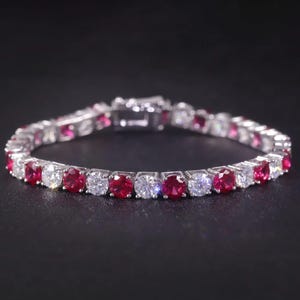 Può includere: Un bracciale d'argento con pietre preziose rotonde alternate rosse e trasparenti. Il bracciale è posto su uno sfondo scuro, evidenziando la brillantezza delle gemme. La chiusura è visibile.