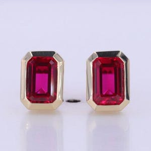 May include: A pair of stud earrings featuring rectangular, emerald-cut vibrant red gemstones set in polished yellow gold bezels. The earrings have a clean, geometric design with a smooth metal frame surrounding each deep red stone.