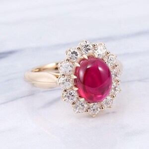May include: A gold ring featuring a large, oval, cabochon-cut deep red gemstone at the centre. The central stone is surrounded by a halo of twelve small, round, clear faceted stones set in a scalloped gold frame with a smooth, polished gold band.
