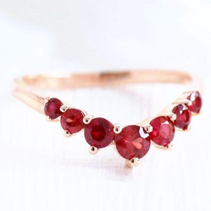 Könnte beinhalten: Ein Ring aus Roségold mit einer geschwungenen, V-förmigen Ringschiene, die mit sieben runden, tiefroten Rubinen besetzt ist. Die Steine sind in abgestufter Größe angeordnet, wobei der größte Rubin in der Mitte der Kurve sitzt und von Metallkrappen gehalten wird.