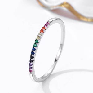 May include: A silver ring with a row of small, colorful gemstones. The gemstones are arranged in a rainbow pattern, with red, orange, yellow, green, blue, indigo, and violet hues. The ring is simple and elegant.