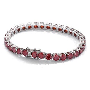 May include: A silver tennis bracelet with a series of round, deep red gemstones. The bracelet has a secure clasp and is designed with a classic, elegant style. The gemstones are evenly spaced and set in a polished metal setting.