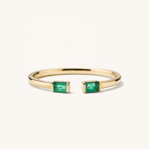 Op de afbeelding: Een open gouden ring met twee smaragdgroene stenen in baguette-slijpvorm aan beide uiteinden van de band. De gepolijste gouden ring heeft een gladde, afgeronde afwerking en de twee rechthoekige groene stenen zitten in eenvoudige metalen zettingen.