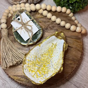 Könnte beinhalten: Zwei dekorative Muschelschalen-Schmuckschalen. Eine ist grün mit goldenem Rand und einem mit Bindfaden befestigten Etikett. Die andere ist weiß mit goldenem Rand und einem goldenen Weihnachtsbaum-Design. Holzperlen und eine Quaste sind ebenfalls sichtbar.