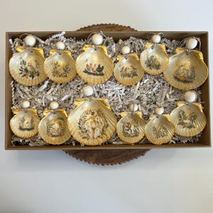 Puede incluir: Un conjunto de adornos de conchas marinas con escenas pintadas, presentados en una caja de madera con relleno de papel. Cada concha tiene un borde dorado, una cuenta blanca y una cinta dorada. Las escenas pintadas incluyen figuras, plantas y animales.