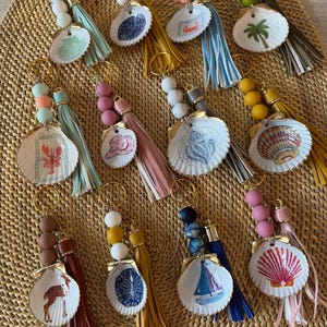 May include: A collection of twelve keychains featuring painted seashells, colourful silicone beads, and leather-like tassels. Each shell displays a unique illustration, including a lobster, palm tree, sailboat, deer, and various coastal motifs.