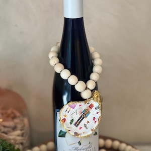 Puede incluir: Una botella de vino azul oscuro con tapa blanca y un adorno decorativo de ostra para vino. El adorno presenta un diseño festivo con un cascanueces y otros elementos navideños. Una guirnalda de cuentas está drapeada alrededor de la botella.