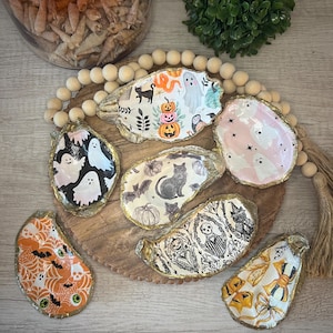 Peut inclure: Une collection de coquilles d'huîtres décoratives sur le thème d'Halloween. Chaque coquille est décorée de motifs effrayants, notamment des fantômes, des citrouilles, des chauves-souris et des chats. Les coquilles ont des bords dorés et sont disposées sur un plateau en bois.