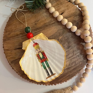 Puede incluir: Adorno navideño de concha marina con un soldado cascanueces pintado. La concha tiene un borde dorado. Está ensartado con un lazo de cuerda y cuentas rojas y verdes. El adorno se exhibe sobre una superficie de madera con cuentas de madera.
