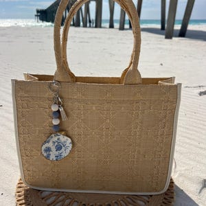 May include: A beige tote bag with a woven texture, white trim, and matching handles. A blue and white floral charm and tassel hang from the handle. The bag sits on a round, woven mat on a sandy beach.