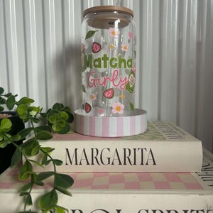 May include: A clear glass tumbler with a bamboo lid and a glass straw. The exterior features a colourful decal with the text "Matcha Girly" in green and pink, surrounded by illustrations of strawberries, pink flowers, leaves, and small stars.