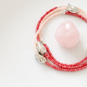 May include: A layered necklace with red and pink beads, silver accents, and a polished rose quartz stone. The necklace features three strands of small beads and abstract silver clasps. The rose quartz is a soft pink color.