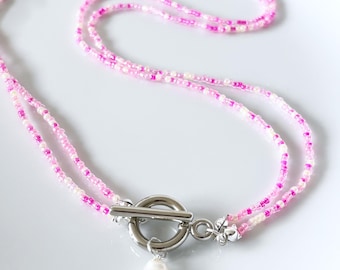 May include: A double-strand necklace featuring small pink and white seed beads. The necklace is secured with a silver-toned metal toggle clasp and features a single white freshwater pearl charm hanging from the center ring of the clasp.