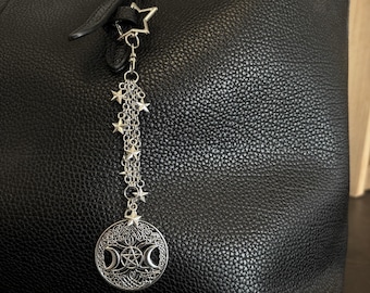 May include: A black textured tote bag with a red interior lining, featuring a silver-toned metal bag charm attached to a black strap with a gold buckle. The charm consists of a chain with small star accents and a circular triple moon pendant.