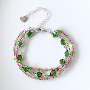 Può includere: Un bracciale a più fili con file alternate di piccole perline color verde lime e rosa acceso. Il design incorpora diverse pietre di vetro rotonde e sfaccettate color verde scuro incastonate in montature argentate, con una chiusura a moschettone.