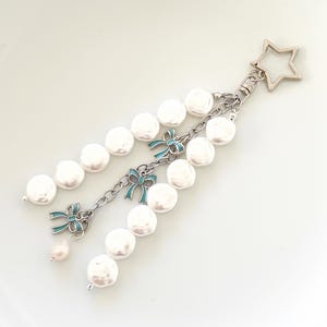 May include: A silver-coloured keychain with a star-shaped clasp. The keychain features a chain with white pearl beads and teal bow accents. A small pearl hangs at the end of the chain. The background is white.