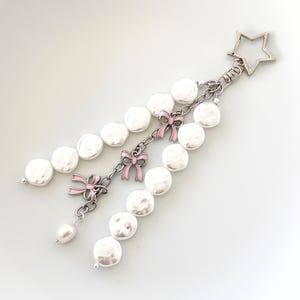 May include: A silver-toned keychain with a star-shaped clasp, featuring a chain of white, flat pearls. Pink enamel bows accent the chain. A single pearl dangles from the bottom. The keychain is on a white background.