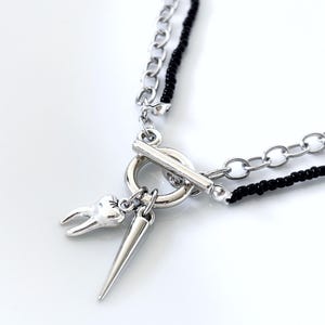 May include: A layered necklace featuring a silver-tone chain and a strand of small black beads. The necklace includes a toggle clasp closure with two dangling charms: one shaped like a human tooth and the other a smooth, pointed silver-tone spike.