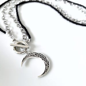 Op de afbeelding: Een meerstrengs ketting met een zilverkleurige halve maan hanger met ingewikkelde geometrische patronen en sterren. De ketting is voorzien van een zilverkleurige knevelsluiting, een metalen ketting en een streng met zwarte kraaltjes.