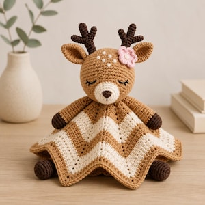 May include: A crochet deer lovey featuring a tan head with dark brown antlers, a white muzzle, and a small pink flower accent. The body is a soft, chevron-patterned blanket in alternating stripes of tan and cream, with dark brown crochet paws.