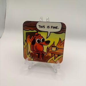 May include: A square coaster featuring a cartoon dog sitting in a burning room, with the text "THIS IS FINE" in a speech bubble. The dog is orange, and the flames are yellow and orange.