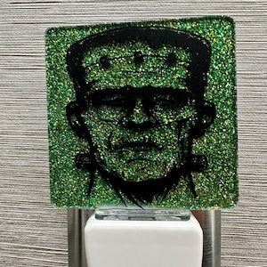May include: A square night light featuring a black silhouette of Frankenstein's monster on a glittery green background. The night light has a white base and is plugged into a silver outlet.