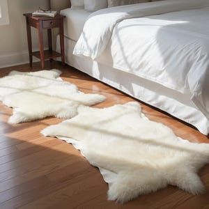 May include: Two off-white sheepskin rugs are placed on a hardwood floor in a bedroom setting. The rugs have an irregular shape and a soft, fluffy texture. The room has a bed with white bedding and a small wooden side table.